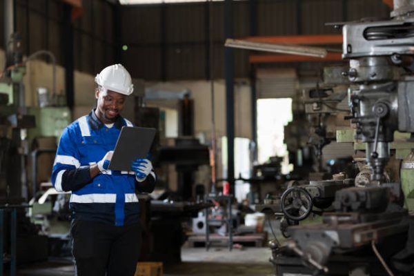 Male engineer worker using digital tablet during working with lathe machine in industry factory, wearing safety uniform, helmet. Male technician worker maintenance parts of machine in workshop plant