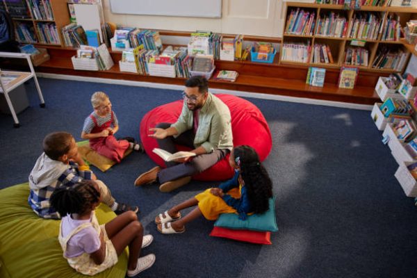 High angle view of enthusiastic male teacher reading a book to primary students while sitting in school library with copy space. Curious elementary schoolchildren sitting on big cushion in library while listening to story from librarian. Top view of middle eastern man at primary school reading fairy tale during recess.